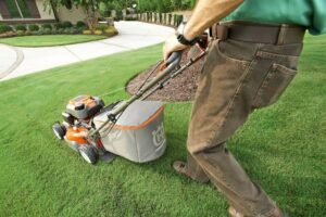 A person wearing a green shirt and brown pants is using an orange and gray lawn mower to mow the green grass