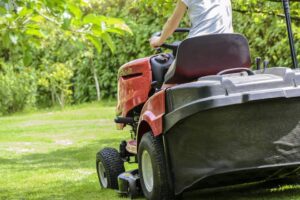 A person wearing a gray shirt is riding a black and orange lawn mower on a green grass
