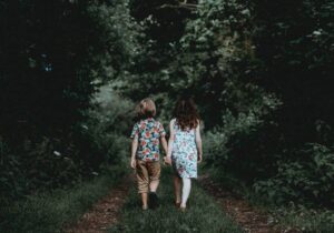 A girl wearing a floral dress walks hand in hand with a boy wearing a floral shirt on green grass inside the forest