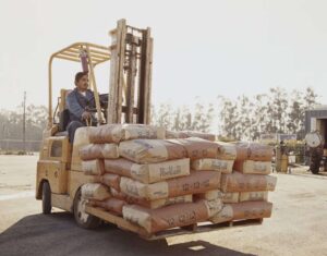 A man wearing a blue coverall suit is moving Milorganite fertilizer with a yellow forklift on a sunny day