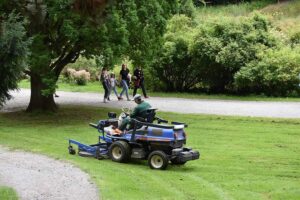 A man wearing green coveralls is riding the blue lawn mower near a group of people walking on a rough walk path