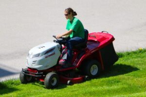 A woman wearing an oversized green shirt and black sunglasses is riding a red and white lawn mower