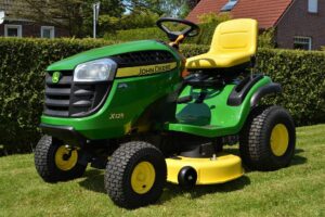A green and yellow mower on green grass near a green hedge